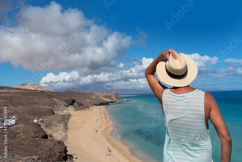 Hombre de espaldas sosteniendo su sombrero de paja frente a la playa de arena blanca y el mar turquesa de Jandia en la localidad turística de Fuerteventura en las Islas Canarias. photo