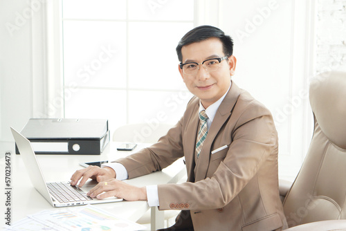 Attractive cheerful asian classy CEO businessman  in suit and with eyeglasses using laptop while looking at the camera, Asian boss middle age. CEO business man in formal clothing wearing spectacles