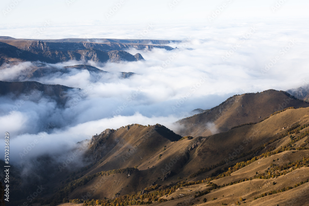 Photo of a scenic view of hills and massive clouds crawling on gorge under sunlight and clouds shadows on the plateau Bermamyt. There are trees in autumn colors on the hills.