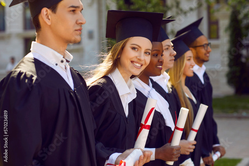 Group of multicultural people in graduation gowns and caps graduate universuty outdoors in campus. They are standing in line. One girl is looking at camera. Graduation from college university concept. photo