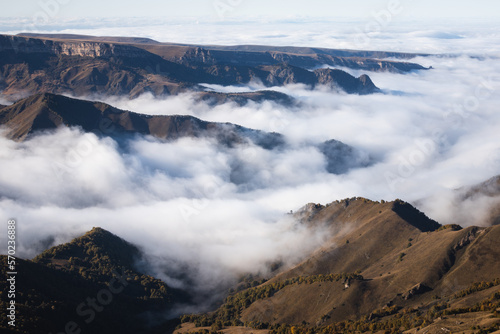 Photo of a scenic view of hills in the lowland and clouds crawling between hills under sunlight and clouds shadows on the plateau Bermamyt. There are trees in autumn colors on the hills.