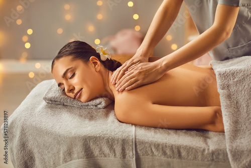 Side view photo of pretty young brunette woman with closed eyes relaxing in spa salon getting massage. Therapist doing manipulative treatment on shoulders. Wellness and beauty day concept