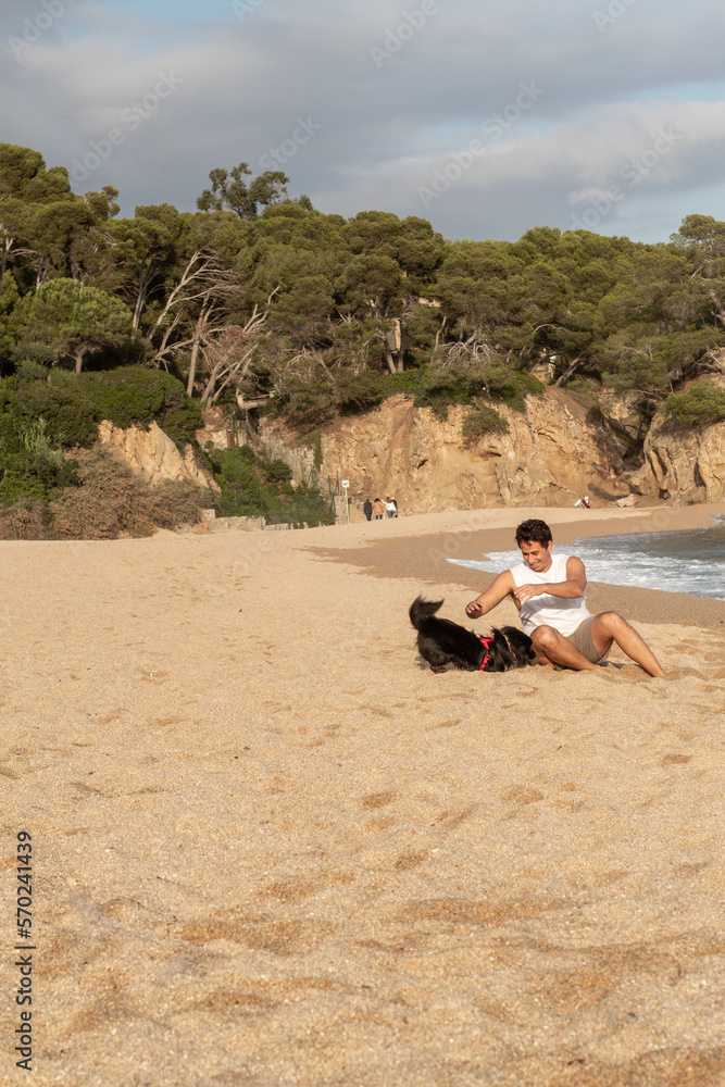 Hombre joven de raza caucásica jugando con su perro negro en la arena de la playa de la costa brava.