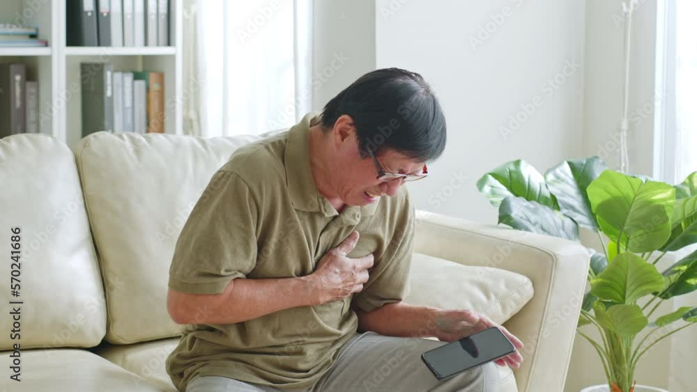 Asian senior man clutch his chest because heart attack symptom during using smartphone at home Asian senior man clutch his chest because heart attack symptom during using smartphone at home