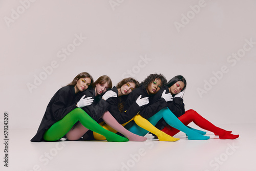 Canvas Print Five young beautiful different women in black jackets and colored tights sitting on floor and looking at camera with lost look over grey background