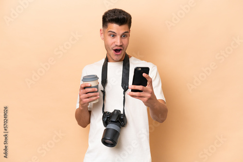 Young photographer caucasian man isolated on beige background holding coffee to take away and a mobile