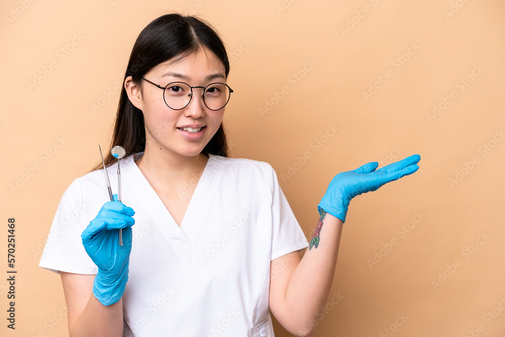 Dentist Chinese woman holding tools isolated on beige background extending hands to the side for inviting to come Dentist Chinese woman holding tools isolated on beige background extending hands to the side for inviting to come