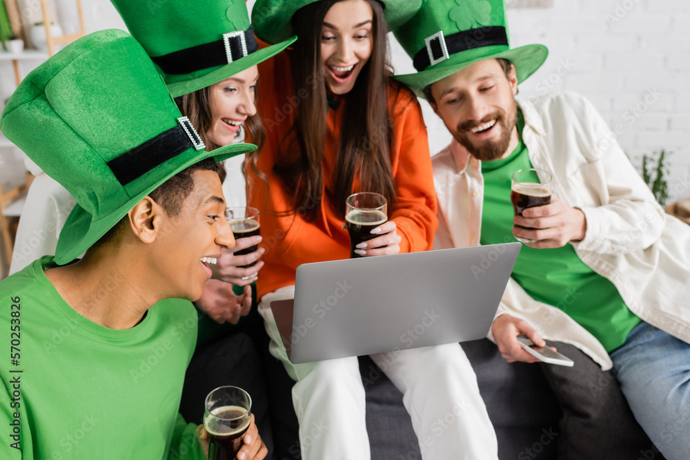 excited interracial friends looking at laptop during video call while holding glasses of dark beer on Saint Patrick Day excited interracial friends looking at laptop during video call while holding glasses of dark beer on Saint Patrick Day