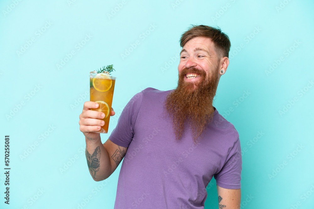 Redhead man with long beard holding a cocktail isolated on blue background with happy expression Redhead man with long beard holding a cocktail isolated on blue background with happy expression