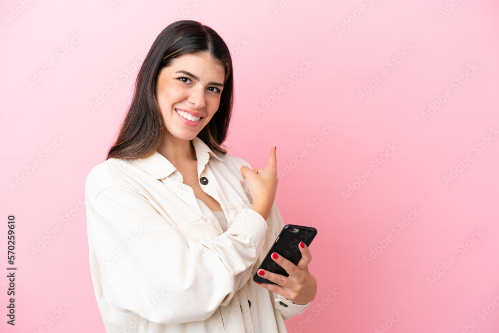 Young Italian woman isolated on pink background using mobile phone and pointing back Young Italian woman isolated on pink background using mobile phone and pointing back