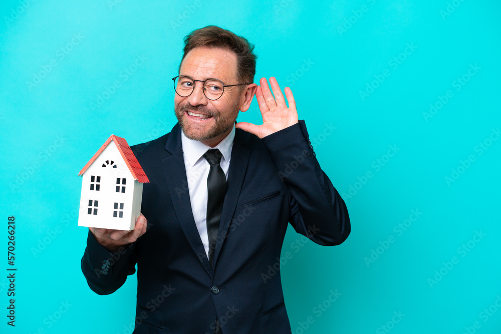 Real estate middle age agent man isolated on blue background listening to something by putting hand on the ear Real estate middle age agent man isolated on blue background listening to something by putting hand on the ear