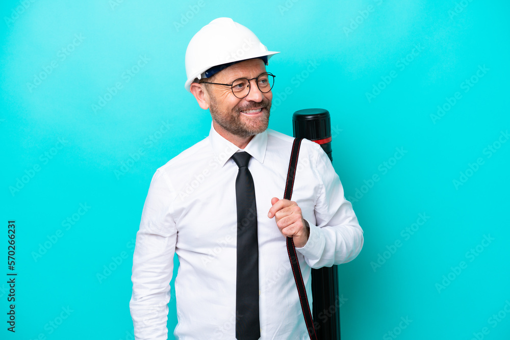 Middle age architect man with helmet and holding blueprints isolated on blue background looking to the side and smiling Middle age architect man with helmet and holding blueprints isolated on blue background looking to the side and smiling