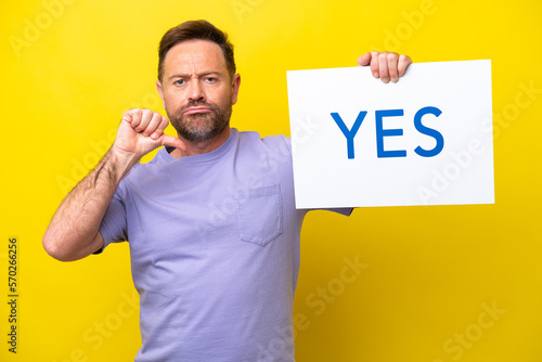 Middle age caucasian man isolated on yellow background holding a placard with text YES with proud gesture © luismolinero