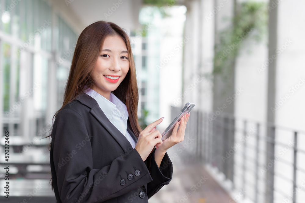 Young professional Asian businesswoman in a white shirt and dark blue suit is standing confidently smiling indoors and looking at the camera while using a smartphone to communicate. Young professional Asian businesswoman in a white shirt and dark blue suit is standing confidently smiling indoors and looking at the camera while using a smartphone to communicate.