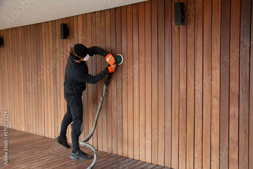 Deck worker in mask sanding hardwood siding with orbital sander, wooden deck and cladding renovation