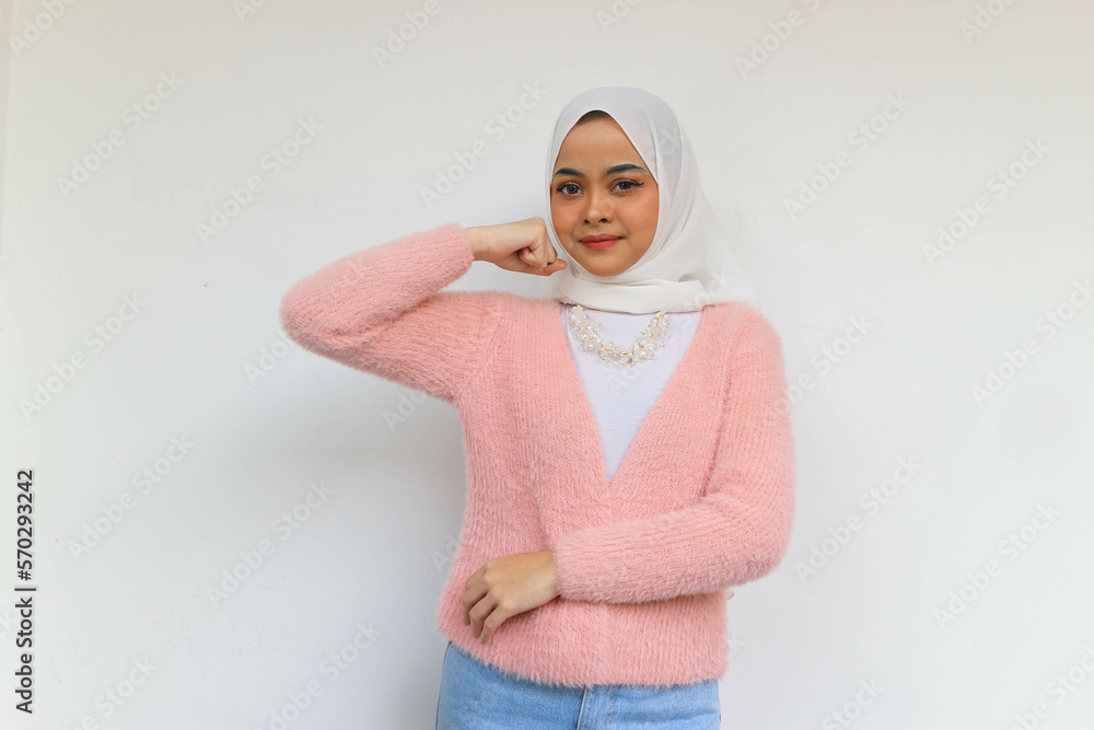 Fototapeta premium Beautiful smiling asian young hijab woman wearing pink blazer, look confident looking at camera while showing the bicep muscle. Isolated on white background