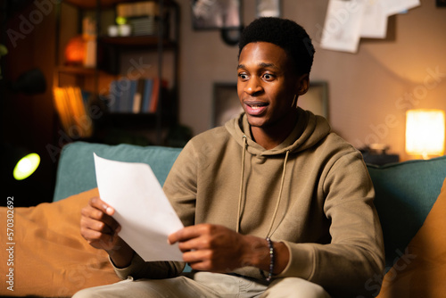 Renting cost time shocked frustrated surprised young african american man in the living room sitting holding documents papers bills in hand reading real estate problem concept.