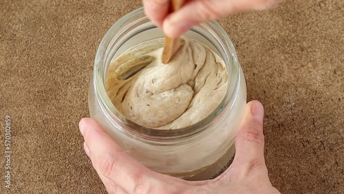 Female hand stirs the wheat sourdough starter with a wooden spoon in a glass jar. Natural yeast for making rye or wheat artisan bread. Fermentation product. Close up. 
