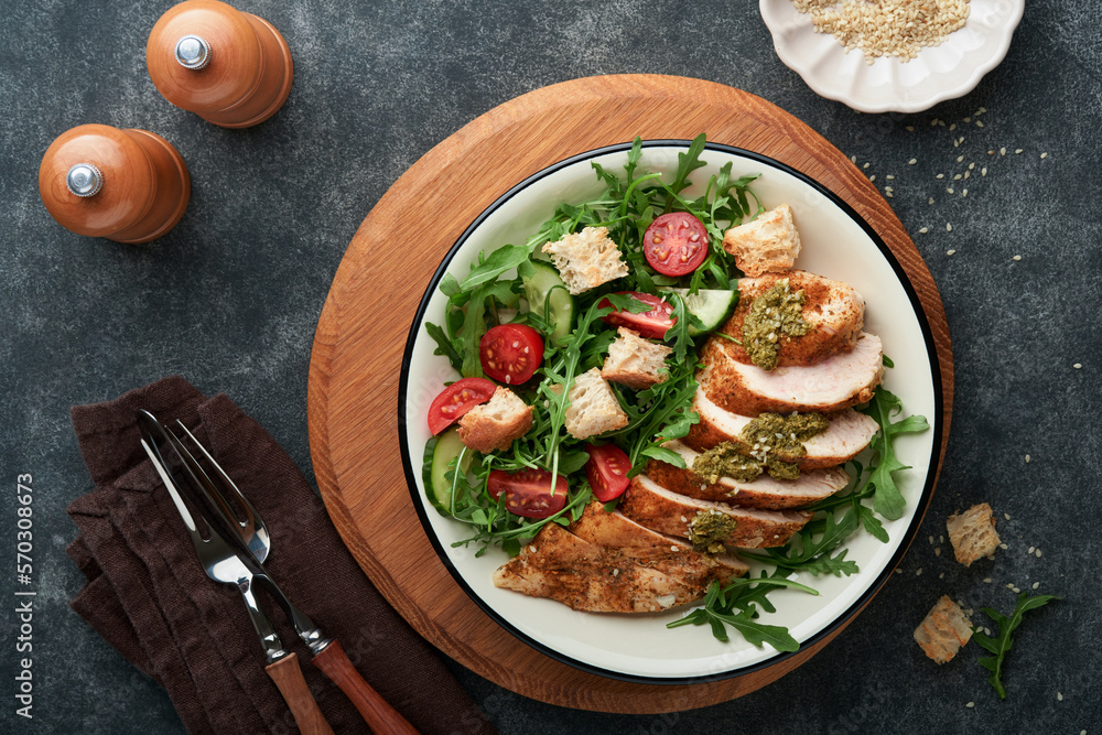 Fresh salad with grilled chicken fillet, lettuce, arugula, tomatoes, cucumber and pesto sauce, sesame seeds with olive oil in white bowl on black slate background. Healthy lunch menu. Top view. Fresh salad with grilled chicken fillet, lettuce, arugula, tomatoes, cucumber and pesto sauce, sesame seeds with olive oil in white bowl on black slate background. Healthy lunch menu. Top view.