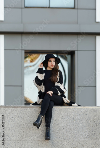 Portrait of a young Caucasian girl sitting on a ledge holding her hat with one hand with a mirrored window building in the background. Vertical lifestyle portrait.