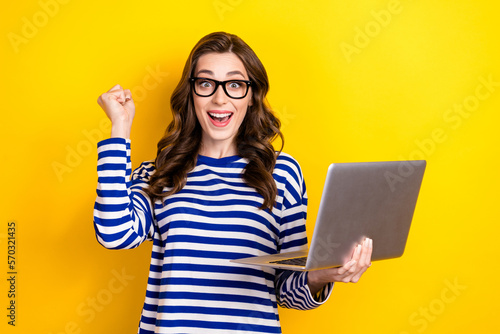 Photo of young excited woman wear new eyeglasses fist up celebrate hold netbook victory online gambling lottery isolated on yellow color background