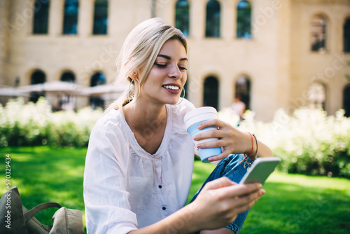 Pretty young female blogger with cellphone gadget enjoying mobile blogging during coffee time in summer park, cheerful hipster girl posting web content publication in social media and smiling photo