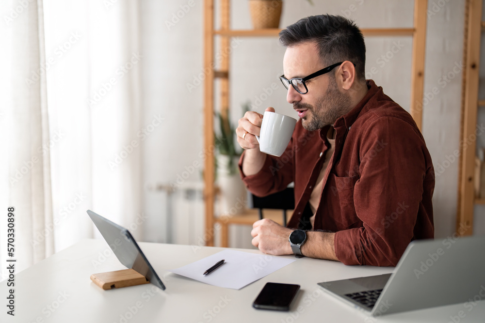 Freelance worker with glasses in casual clothing holding cup, drinking coffee in home office, sitting near window. Laptop computer, digital tablet device, smart phone, paper document and pen on table. Freelance worker with glasses in casual clothing holding cup, drinking coffee in home office, sitting near window. Laptop computer, digital tablet device, smart phone, paper document and pen on table.