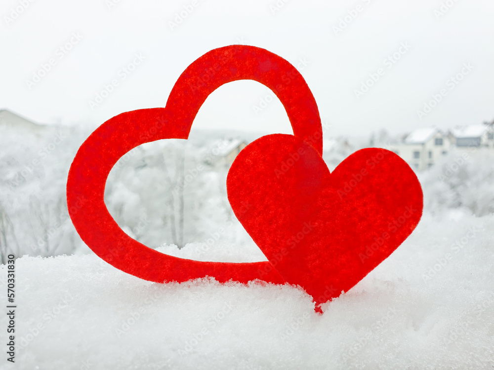 Two hearts cut out of red felt is standing in the snow. Heart-shaped hole is cut in bigger heart. Blurred background of cottage village in winter. Valentine's day concept.