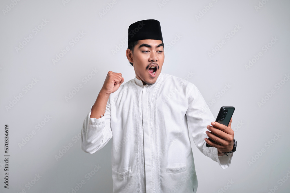Asian muslim man in white clothes holding smartphone doing winning gesture holding mobile phone Asian muslim man in white clothes holding smartphone doing winning gesture holding mobile phone