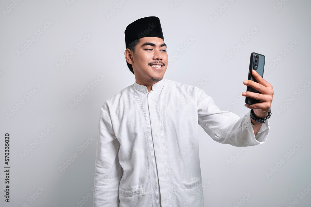 Cheerful young Asian Muslim man doing video call or selfie shot on mobile phone Cheerful young Asian Muslim man doing video call or selfie shot on mobile phone