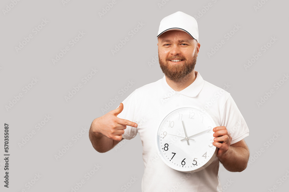 Cheerful delivery man wearing white points at a round clock he is holding. Studio shot over grey background.