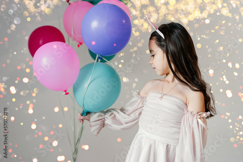 Little beautiful shy brunette girl wearing a party hat and pink tutu dress holding balloons in a party. Portrait little pretty child with blue, green and pink balloons.