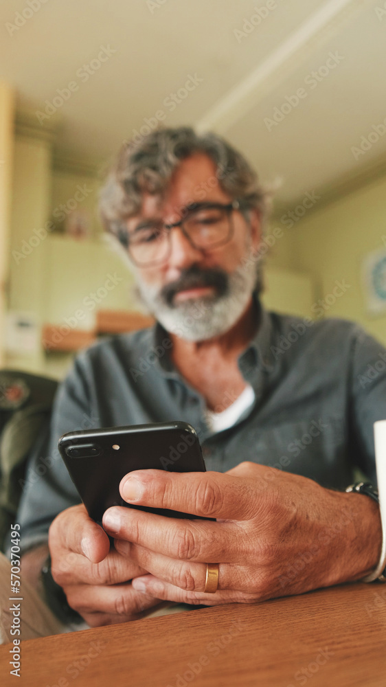 Close-up of an elderly man attentively reading message on his smartphone and drinking coffee while sitting in the kitchen Close-up of an elderly man attentively reading message on his smartphone and drinking coffee while sitting in the kitchen