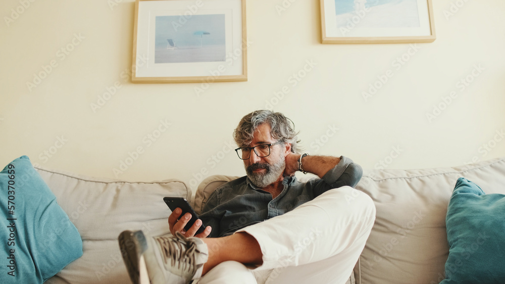 Middle-aged man relaxing on his couch while in home and using his smartphone to text Middle-aged man relaxing on his couch while in home and using his smartphone to text