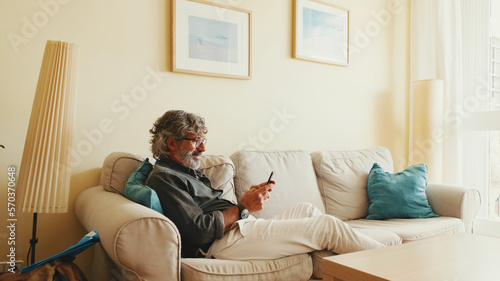 Middle-aged man relaxing on his couch while in home and using his smartphone to text © Andrii Nekrasov