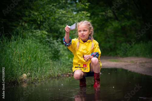 A blonde girl with pigtails in a yellow raincoat and boots launches a paper boat in a puddle in summer