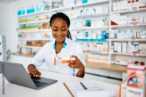 Happy African American pharmacist working on laptop while analyzing medicine in pharmacy.