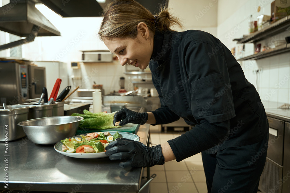 Smiling female chef in restaurant kitchen decorates plate with salad Smiling female chef in restaurant kitchen decorates plate with salad