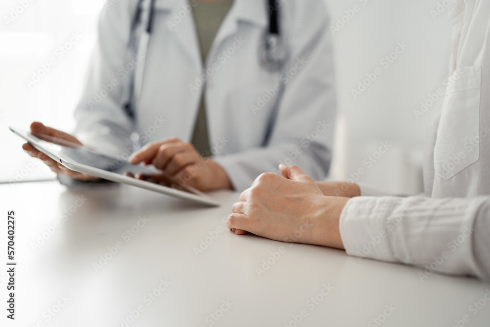 Doctor and patient sitting at the desk in clinic office. The focus is on female physician's hands using tablet computer, close up. Perfect medical service and medicine concept Doctor and patient sitting at the desk in clinic office. The focus is on female physician's hands using tablet computer, close up. Perfect medical service and medicine concept