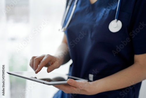 Doctor woman using tablet computer while standing near panorama window in clinic, close up. Physician or surgeon at work. Medicine concept photo