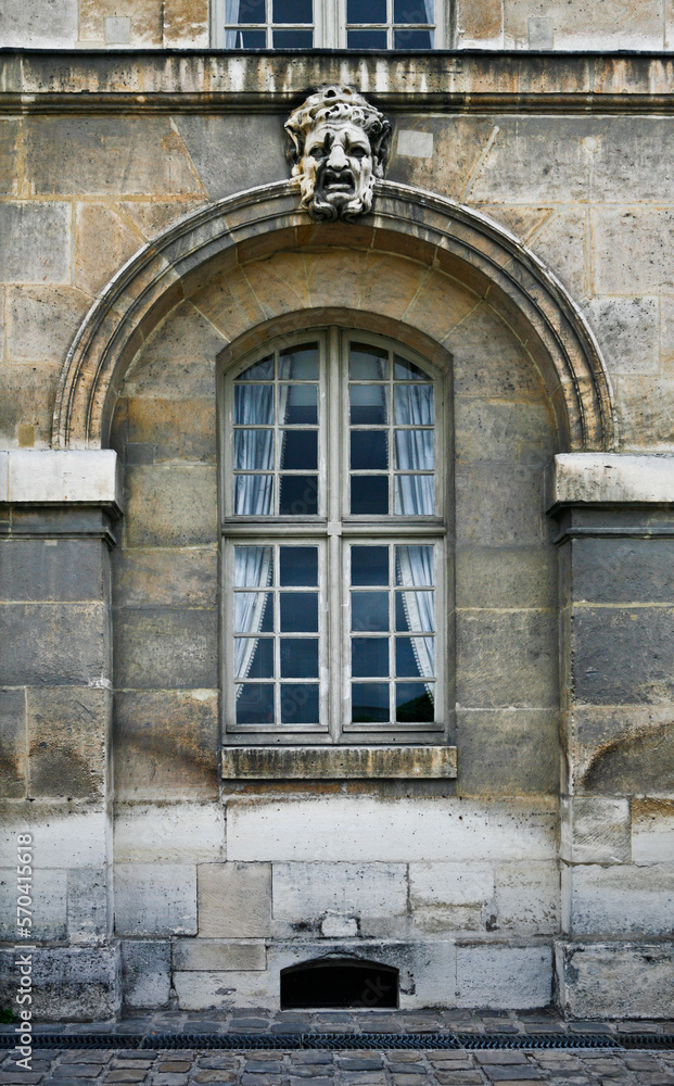 Obraz premium Paris ancient stone building facade with a French window, white curtains, stucco fretwork arch and dramatic mask, and cobblestone pavement