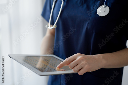 Doctor woman using tablet computer while standing near panorama window in clinic, close up. Physician or surgeon at work. Medicine concept photo