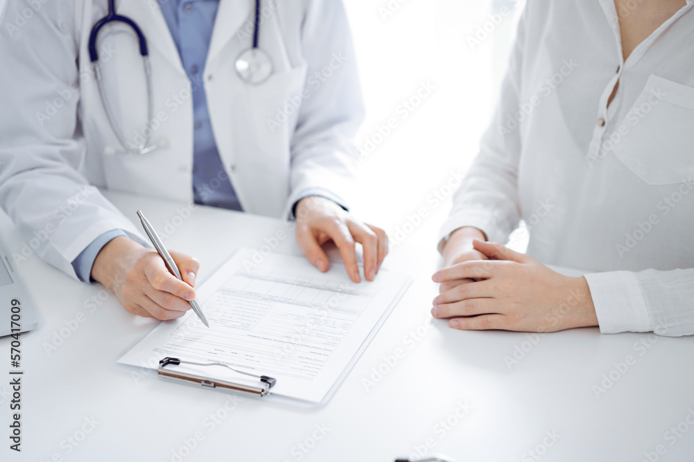 Doctor and patient sitting near of each other at the desk in clinic. The focus is on female physician's hands filling up the medication history record form or checklist, close up. Medicine concept Doctor and patient sitting near of each other at the desk in clinic. The focus is on female physician's hands filling up the medication history record form or checklist, close up. Medicine concept