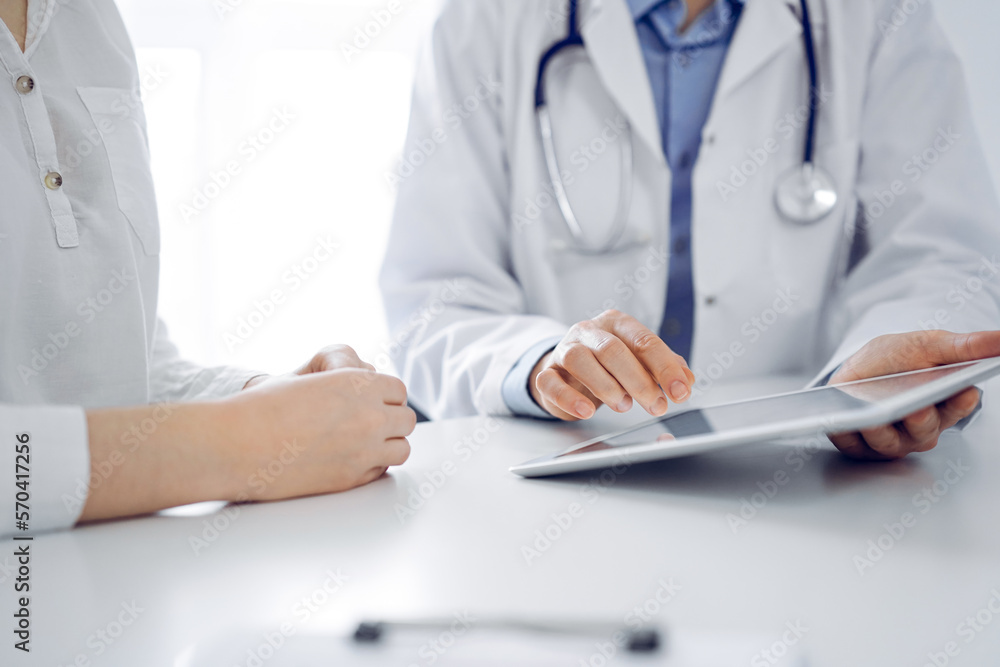 Doctor and patient sitting near each other at the desk in clinic. The focus is on female physician's hands pointing into tablet computer touchpad, close up. Medicine concept Doctor and patient sitting near each other at the desk in clinic. The focus is on female physician's hands pointing into tablet computer touchpad, close up. Medicine concept