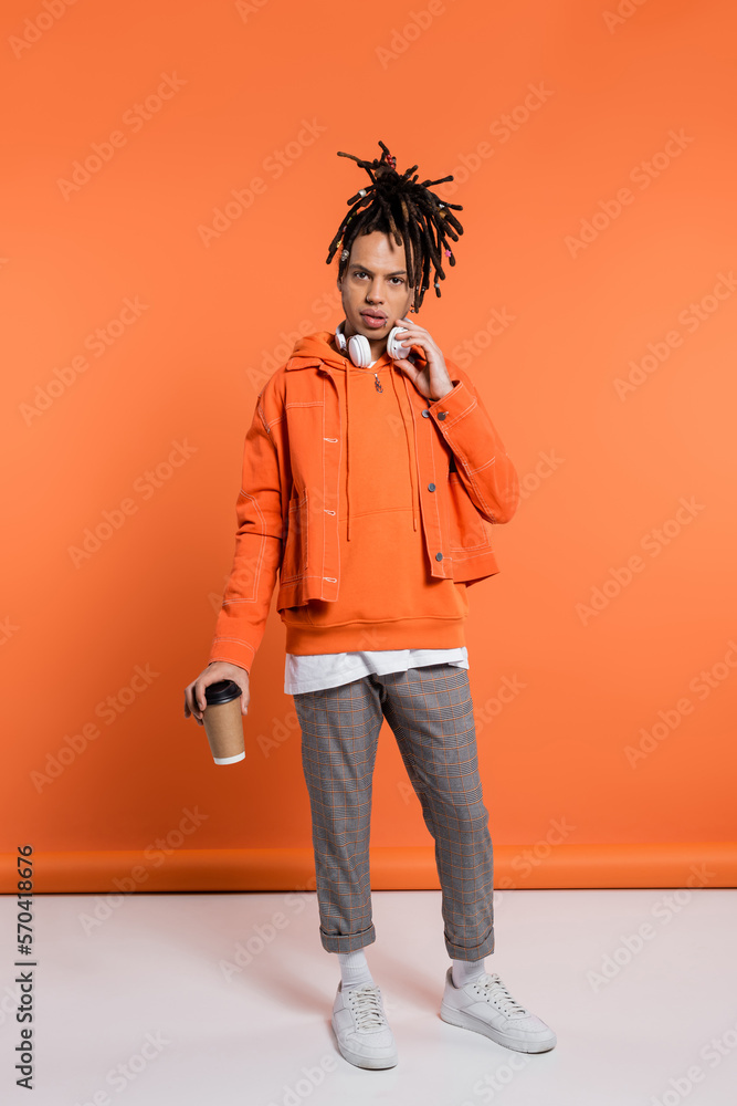 Fototapeta premium pierced and multiracial man with wireless headphones holding paper cup with coffee to go on coral background