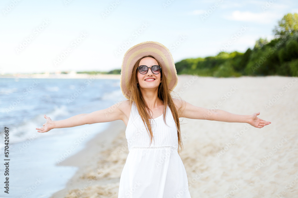 Happy smiling woman in free happiness bliss on ocean beach standing with a hat, sunglasses, and raising hands. Portrait of a multicultural female model in white summer dress enjoying Happy smiling woman in free happiness bliss on ocean beach standing with a hat, sunglasses, and raising hands. Portrait of a multicultural female model in white summer dress enjoying