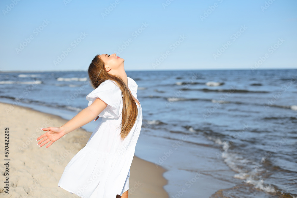 Happy, beautiful woman on the ocean beach standing in a white summer dress, open arms