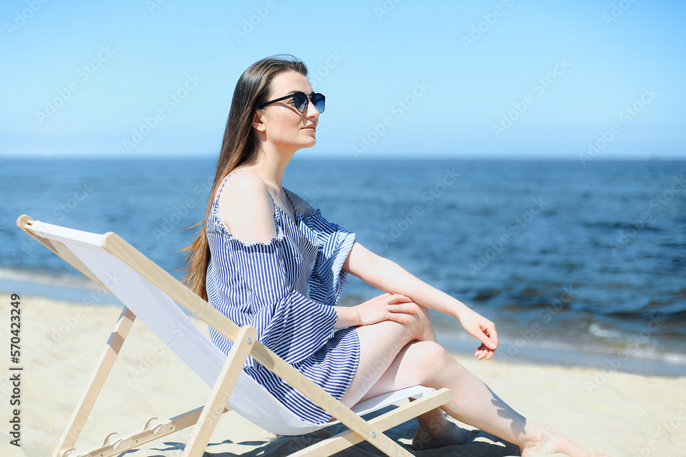 Happy young brunette woman relaxing on a wooden deck chair at the ocean beach while smiling, and wearing fashion sunglasses. The enjoying vacation concept