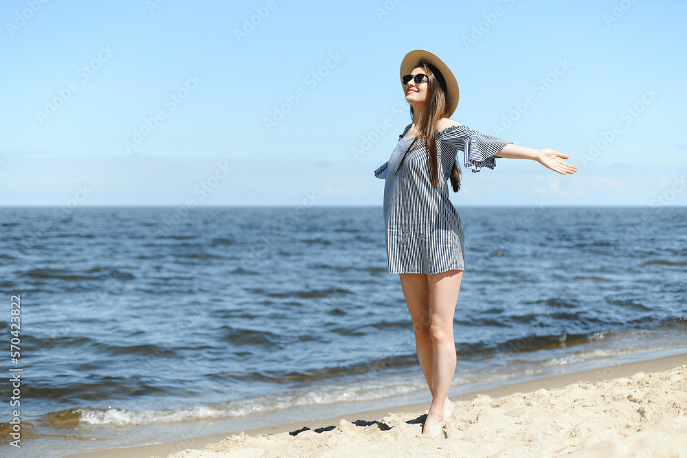 Happy smiling woman in free bliss on ocean beach standing with open hands. Brunette female model in sunglasses and hat enjoying nature during travel holidays vacation outdoors, medium shot