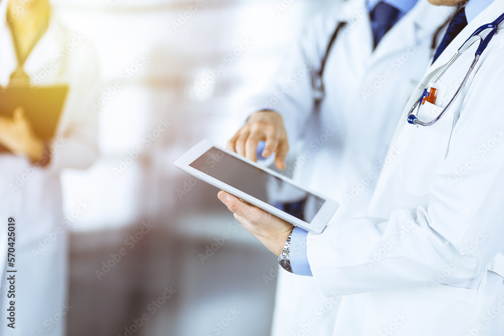 Group of unknown doctors use a computer tablet to check up some medical names records, while standing in a sunny hospital office. Physicians ready to examine and help patients Group of unknown doctors use a computer tablet to check up some medical names records, while standing in a sunny hospital office. Physicians ready to examine and help patients